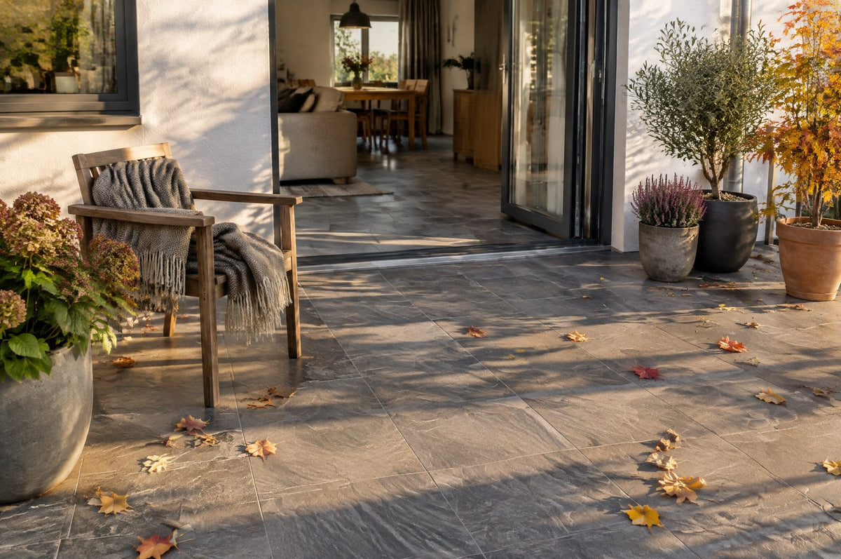 Terrasse mit großformatigen Platten in grauer Steinoptik, Gartenstuhl mit Decke und Herbstlaub, Blick ins Haus